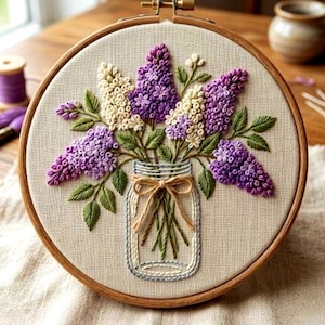 Peut inclure: Broderie florale sur cerceau, représentant un bouquet de lilas violets et blancs dans un bocal en verre. La broderie est réalisée sur un tissu beige, encadrée par un cerceau en bois. Des feuilles vertes et un nœud beige complètent le motif.