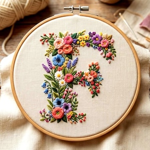 Könnte beinhalten: Ein floraler Stickrahmen mit dem Buchstaben "F", verziert mit bunten Blumen und Grünzeug. Die Stickerei zeigt rosa, blaue, gelbe und violette Blüten mit grünen Blättern und Stielen. Der Rahmen ist aus Holz, der Stoff ist hellbeige.