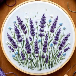 Puede incluir: Un bastidor de bordado terminado con un diseño de campo de lavanda. El diseño incluye lavanda morada, flores blancas y azules y tallos verdes. Hilos de bordar y herramientas, incluyendo tijeras y agujas, rodean el bastidor sobre una superficie de madera.