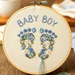 May include: Embroidered hoop art with the words "BABY BOY" in blue thread. The design features floral footprints in blue, white, and green. The hoop is set against a wooden surface, with a cup of tea in the background.
