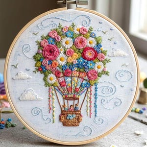 May include: Embroidered hot air balloon hoop art featuring a basket with passengers, suspended beneath a vibrant bouquet of flowers. The flowers include pink roses, white daisies, and blue blossoms, set against a light blue background with embroidered clouds and birds.