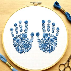 May include: A hand-stitched embroidery hoop featuring a floral design in shades of blue. The design depicts two hands filled with flowers and leaves. Embroidery floss and small scissors are also visible.