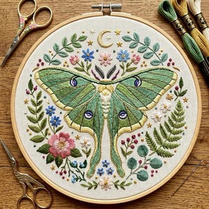 May include: An embroidery hoop containing a fabric panel with a central green luna moth surrounded by embroidered flowers, leaves, and a crescent moon. Two pairs of gold-toned scissors and three skeins of embroidery floss are positioned nearby.