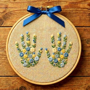 Op de afbeelding: Geborduurd handvormig ontwerp met blauwe, gele en witte bloemen op beige stof, geplaatst in een houten borduurring. Een blauw lint is bovenaan vastgebonden. Het kunstwerk wordt gepresenteerd op een houten oppervlak.