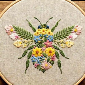 May include: A floral embroidered bee design on a beige fabric within a wooden hoop. The bee is made with yellow, blue, pink, and white flowers, green leaves, and antennae. A handmade embroidery piece.