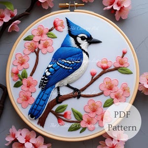 May include: A detailed embroidery hoop art piece featuring a blue jay perched on a branch with pink cherry blossoms. The bird has blue, white, and black feathers. The embroidery is set in a wooden hoop. The text "PDF Pattern" is visible.