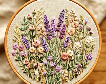 May include: A detailed floral embroidery in a wooden hoop. The design features a variety of flowers in shades of purple, pink, peach, and white, with green leaves and stems. The fabric is a neutral beige color, and the hoop is a light brown.