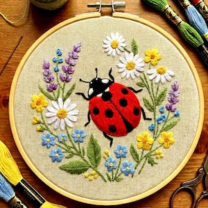 May include: Embroidered hoop art featuring a red ladybug with black spots, surrounded by colorful flowers and greenery. The flowers include white daisies, yellow and blue blossoms, and purple lavender. The embroidery is set in a wooden hoop.