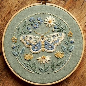 Può includere: Opera d'arte ricamata in un cerchio di legno. Il design presenta una farfalla dettagliata con ali blu e crema, circondata da fiori blu e gialli e foglie verdi, il tutto su un tessuto verde salvia.