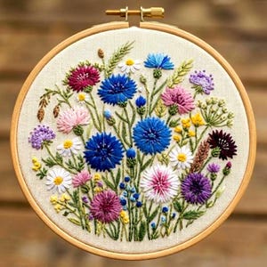 以下が含まれることがあります： カラフルな花の刺繍デザインが施された丸い刺繍枠。刺繍された花は青、ピンク、紫、白の色合いで、緑の茎と葉があります。枠は明るい色の木材で作られています。
