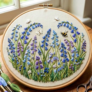 May include: A hand-stitched embroidery piece in a wooden hoop. The design depicts bluebell flowers in shades of blue and purple, with green stems and leaves. Butterflies and a bee are also embroidered. The background is a light beige fabric.