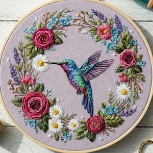 May include: Embroidered hummingbird art in a wooden hoop. The bird is depicted with vibrant blue, green, and purple feathers, surrounded by a floral wreath of pink roses, daisies, and blue flowers. A thimble and scissors are visible.
