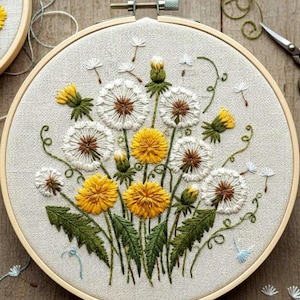 May include: Embroidered dandelion design in a wooden hoop. The artwork features white, yellow, and green thread, depicting dandelions in various stages of bloom. The background is a light beige fabric, and the hoop is made of natural wood.