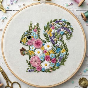Puede incluir: Diseño de ardilla bordada en un aro de madera. La ardilla está llena de flores coloridas, incluyendo rosas rosas, margaritas blancas y flores azules, sosteniendo una bellota. El fondo es una tela color crema.