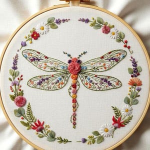 May include: Embroidered dragonfly design in a wooden hoop. The dragonfly has detailed wings with floral patterns in various colours. A floral wreath of red, pink, and white flowers surrounds the dragonfly on a white fabric background.