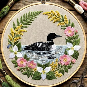 May include: Embroidered artwork of a loon in water, encircled by a floral wreath. The wreath includes green ferns, yellow flowers, pink and white blossoms, and green leaves. The embroidery is set in a wooden hoop.