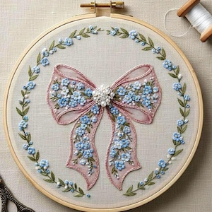 May include: Embroidered artwork featuring a pink bow adorned with blue flowers and a pearl-like centerpiece. The bow is surrounded by a wreath of green leaves and blue flowers, all set within a wooden embroidery hoop. A spool of thread is visible.