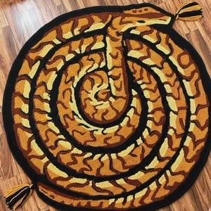 Op de afbeelding: Een ronde, handgetufte vloerkleed met een slangenmotief. Het kleed heeft een zwarte rand en een opgerold slangenpatroon in bruin, beige en geel. Kwasten in zwart, geel en beige zijn bevestigd. Het kleed ligt op een houten vloer.