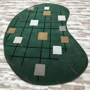 Könnte beinhalten: Ein grüner, nierenförmiger Teppich mit einem Gittermuster aus schwarzen Linien und verstreuten Quadraten in Weiß, Beige und Grau. Der Teppich hat eine weiche Textur und ein modernes, geometrisches Design, geeignet für ein Wohn- oder Schlafzimmer. Maße in Zentimetern.