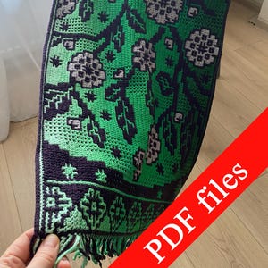 May include: A green and purple crocheted table runner with a floral pattern. The runner features a repeating design of stylized flowers and leaves in shades of green, purple, and white. The edges are finished with a fringe.