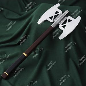 May include: A double-bladed axe with a silver head and a dark brown wooden handle. The axe head features a geometric design. The handle has a black grip with gold accents. The axe is displayed on a dark green fabric.
