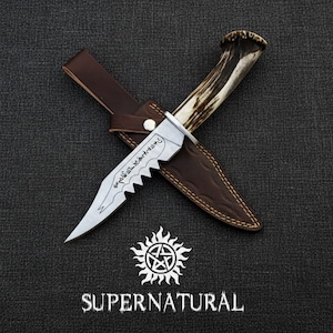 Könnte beinhalten: Ein silbernes Messer mit gezackter Klinge und geätzten Details, das auf einer braunen Lederscheide liegt. Der Griff ist aus Geweih. Das Wort "SUPERNATURAL" steht darunter, mit einem Sonnen- und Sternsymbol.