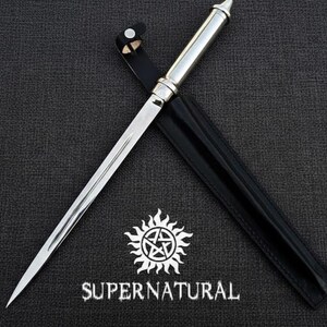 Pu&ograve; includere: Un pugnale d'argento con una lama appuntita e un manico decorativo, in un fodero di pelle nera. La parola "SUPERNATURAL" &egrave; stampata sotto un simbolo di sole bianco con un pentagramma.