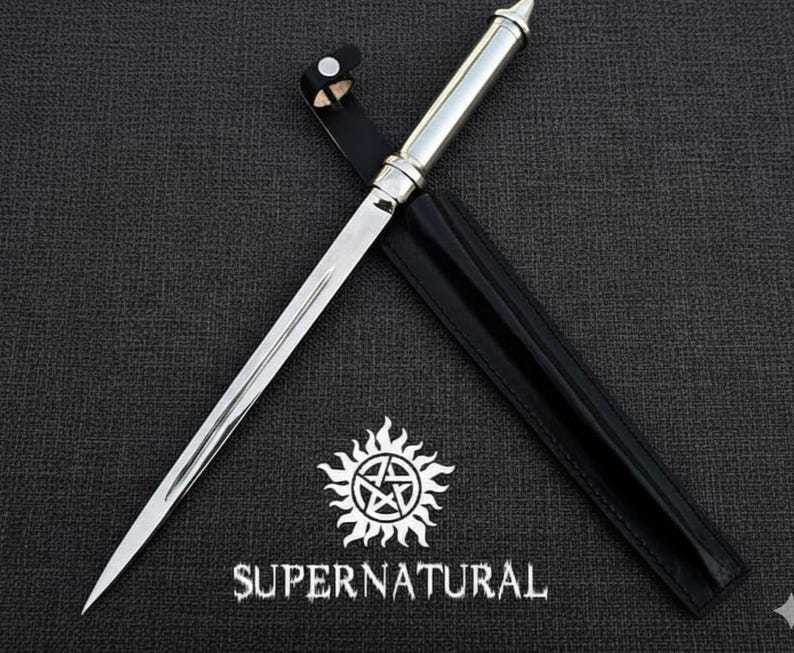 Pu&ograve; includere: Un pugnale d'argento con una lama appuntita e un manico decorativo, in un fodero di pelle nera. La parola "SUPERNATURAL" &egrave; stampata sotto un simbolo di sole bianco con un pentagramma.