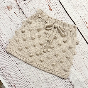 May include: A beige knitted skirt with a textured, bobble design. The skirt features a drawstring waist and a soft, neutral color, perfect for a variety of outfits. The skirt is displayed on a light-colored wooden surface.