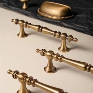 Op de afbeelding: Antieke messing kastbeslagset, inclusief handgrepen, een knop en een greep. De handgrepen hebben een gedetailleerd, sierlijk ontwerp met een klassieke esthetiek. Het beslag wordt gepresenteerd op een neutrale achtergrond, die de warme, metallic afwerking benadrukt.