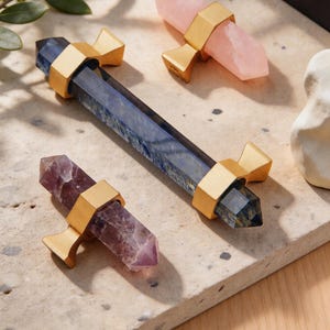 以下が含まれることがあります： ピンク、紫、青色のクリスタルワンドのコレクション。それぞれ金色の金属バンドで固定されています。クリスタルは明るい色のテクスチャ加工された表面に配置されており、自然な要素と装飾に焦点を当てていることを示唆しています。
