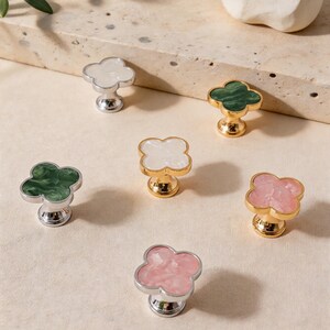 Puede incluir: Una colección de tiradores decorativos para armarios con diseño de trébol. Los tiradores presentan varios colores, incluyendo blanco, verde y rosa, con bases doradas y plateadas. Los tiradores están hechos de un material brillante y están dispuestos sobre una superficie neutra.