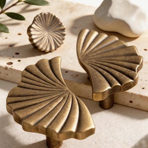 May include: A collection of antique brass cabinet hardware. The set includes two fan-shaped pulls, a round knob, and a small, white stone. The hardware has a textured, aged finish, and is displayed on a neutral surface.