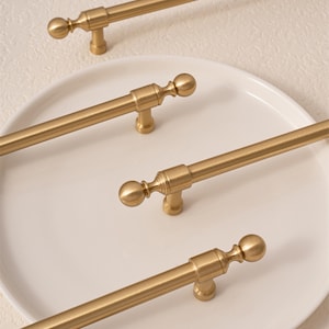 Peut inclure: Quatre poignées de meubles dorées au design classique. Chaque poignée présente une longue barre droite avec un fleuron décoratif à chaque extrémité et une petite base ornée. Les poignées sont disposées sur une surface blanche.