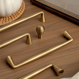 May include: A selection of antique brass cabinet handles and a knob. The hardware has a warm, golden colour and a classic design. The handles are of various sizes and shapes, suitable for drawers and cabinets.