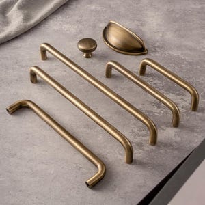 May include: A selection of antique brass cabinet hardware, including bar pulls, a knob, and a cup pull. The hardware is arranged on a grey surface. The text "Prprfctn Treasures" is visible in the top left corner.