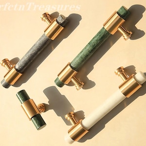 Puede incluir: Cuatro tiradores de armario con mangos cilíndricos de mármol gris, verde y blanco, acentuados con herrajes metálicos dorados. Los tiradores están dispuestos en diagonal sobre una superficie beige claro, mostrando su diseño y materiales.