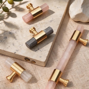 Könnte beinhalten: Schrankbeschläge mit goldfarbenen Metallakzenten und edelsteinartigen Mittelstücken. Die Beschläge umfassen rosa, graue und klare Elemente in rechteckiger und zylindrischer Form. Präsentiert auf einer neutralen Oberfläche.