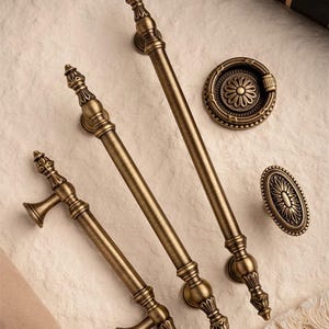 Może przedstawiać: Zestaw antycznych mosiężnych okuć do szafek, w tym dwa długie uchwyty, mniejszy uchwyt, okrągła gałka i owalna gałka. Okucia charakteryzują się bogatymi detalami i estetyką vintage. Widoczny jest tekst "Prprfctn Treasures".