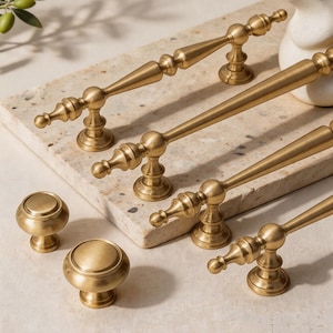 May include: A collection of gold-toned cabinet hardware, including handles and knobs. The handles have a classic design with a central sphere and tapered ends. The knobs are round with a raised, circular top. The hardware is arranged on a light-colored surface.