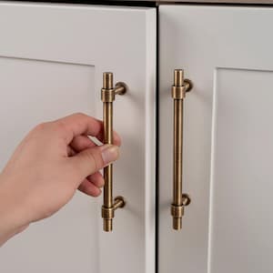 May include: Bronze-colored cabinet pulls with a vertical bar design. The pulls are mounted on white cabinet doors. One hand is shown holding one of the pulls. The pulls have a vintage or antique aesthetic.