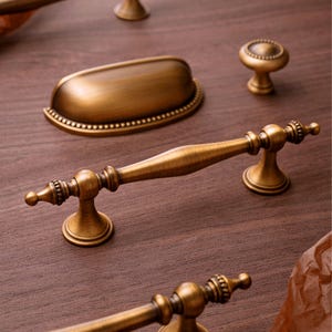 May include: Antique brass cabinet hardware set, including a pull handle, a cup pull, and a knob. The hardware has a detailed, ornate design with a warm, aged finish. The pieces are arranged on a dark wood surface.