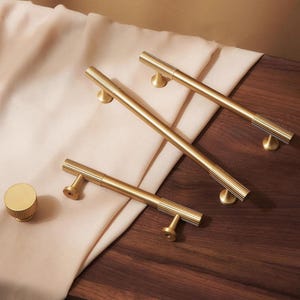 May include: A collection of gold-toned cabinet hardware, including three long bar pulls and a round knob. The pulls have a ribbed design on the ends and are mounted on a dark wood surface with a beige fabric.