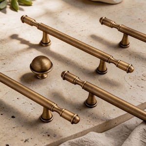 May include: Bronze-toned cabinet hardware set, including several bar pulls and a round knob. The pulls have a classic design with decorative detailing and are arranged on a light-colored surface. The hardware is likely for kitchen or bathroom cabinets.