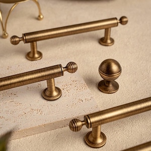 May include: Bronze-toned cabinet hardware set. Includes several bar pulls with ribbed ball ends and a round knob. The hardware has a vintage aesthetic and is displayed on a neutral surface. The pulls are approximately 6 inches long.