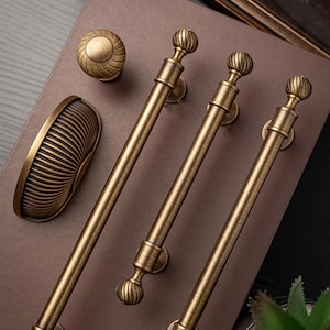 May include: An assortment of antique brass cabinet hardware, including three long pull handles, a round knob, and a curved handle. The hardware has a detailed, ribbed design and a warm, aged finish, suggesting a vintage aesthetic.