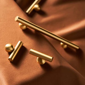 May include: Close-up of several gold-colored cabinet pulls. The pulls have a cylindrical bar design with a spherical accent on each end. The hardware is set against a brown, textured background, creating a warm and inviting aesthetic.