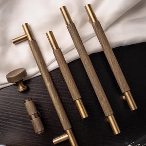 May include: A collection of brass cabinet hardware, including four textured bar pulls, a round knob, and a cylindrical pull. The hardware has a warm, antique gold finish and is arranged on a dark wood surface with a white fabric backdrop.