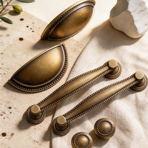 May include: Antique brass cabinet hardware set. The set includes two cup pulls, two bar pulls, and two round knobs. Each piece features a beaded edge design and a warm, aged finish, perfect for adding a vintage touch to furniture.