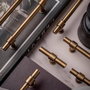 Może przedstawiać: Zbliżenie na kilka antycznych mosiężnych uchwytów i gałek do szafek. Okucia mają cylindryczną konstrukcję z centralnym pasem i są ułożone na marmurowej i ciemnej powierzchni. Uchwyty mają ciepły, metaliczny kolor.